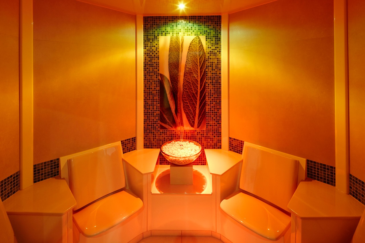 Bowl of white stones rests on a pedestal, centered between two molded benches, as warm orange lights bathe a tiled alcove with large leaf artwork, creating a symmetrical spa-like room.